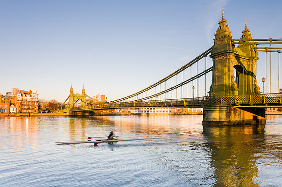 A sculler on the River Thames passes underneath the iron suspension bridge of Hammersmith Bridge, London, in the late afternoon sunshine.